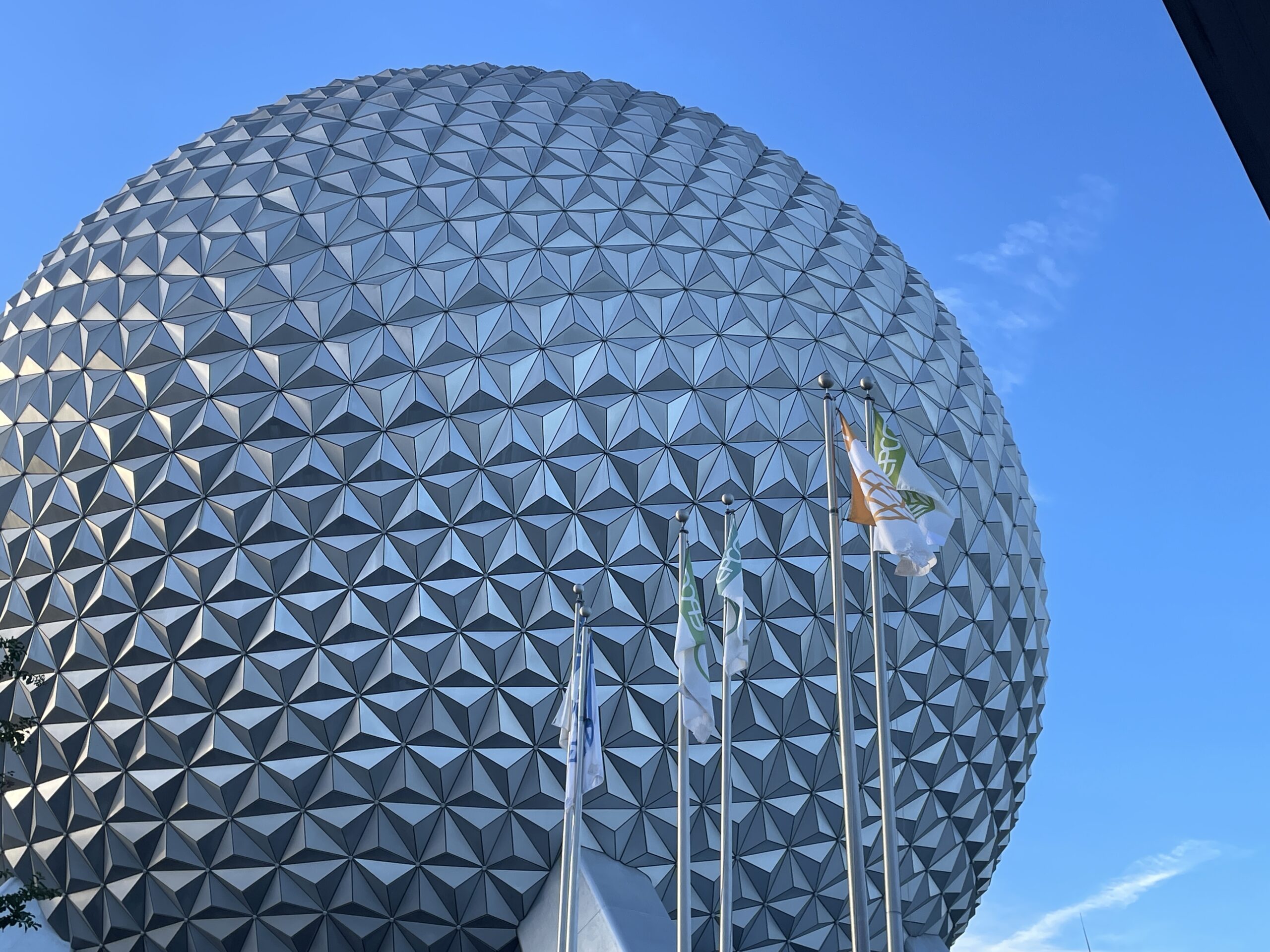 A wide-angle, eye-level view of the iconic Spaceship Earth geosphere at EPCOT. The massive silver structure stands tall against a clear blue sky, framed by manicured green gardens and modern architectural lines. In the foreground, the clean walkways of World Celebration lead the eye toward the attraction, capturing the futuristic and optimistic spirit of the park.