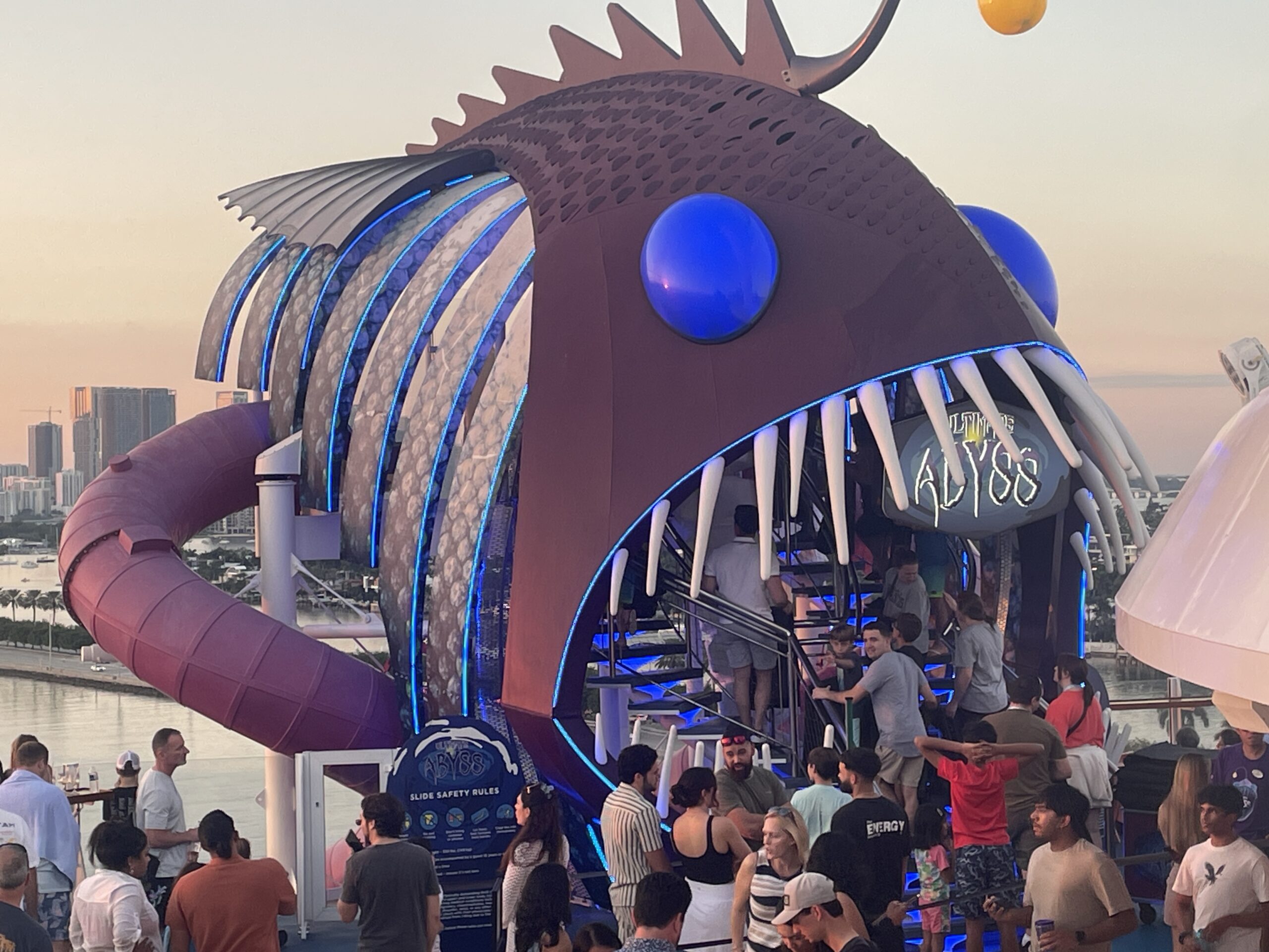 A stunning wide-angle shot of the Wonder of the Seas cruise ship at sea. The image shows the vibrant top decks, including the colorful water slides of the Perfect Storm and the towering Ultimate Abyss slide. The deep blue ocean stretches to the horizon under a sunny sky, highlighting the ship's massive scale and the various outdoor neighborhoods like the Boardwalk and Central Park.