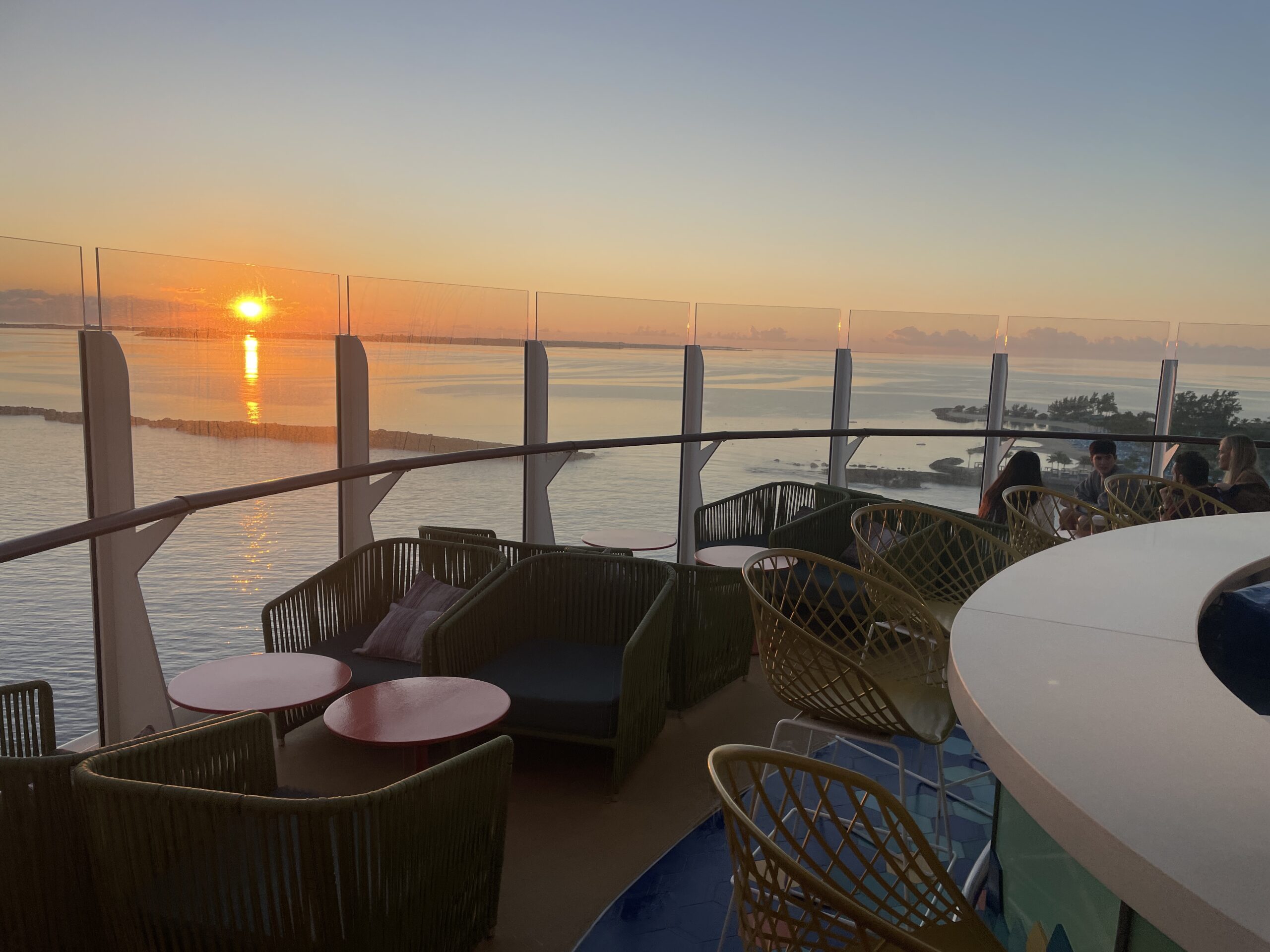 A peaceful orange and gold sunrise over the calm ocean as seen from the wooden railing of a luxury cruise ship deck while enjoying a relaxing vacation.