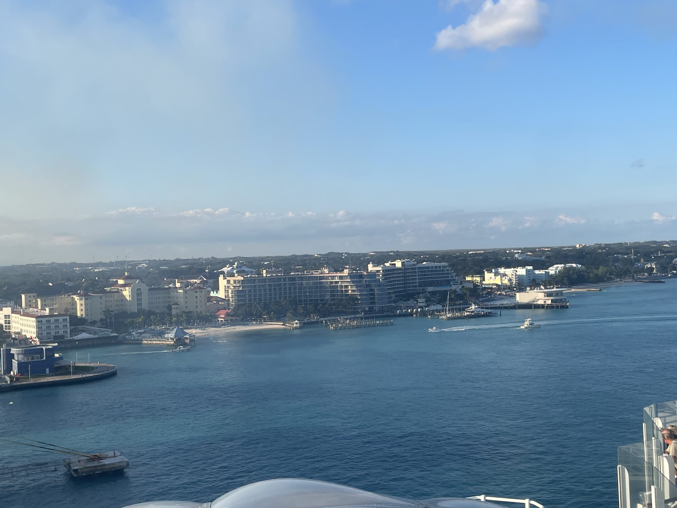 Panoramic view of Nassau Harbor from a cruise ship, featuring several large vessels docked at the upgraded berths, the vibrant pink and pastel colonial buildings of downtown Nassau in the background, and clear Caribbean waters