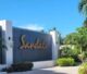 A Sandals Resorts welcome sign surrounded by tropical hibiscus flowers and palm trees at the entrance of a luxury Jamaican all-inclusive resort.