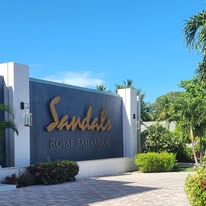 A Sandals Resorts welcome sign surrounded by tropical hibiscus flowers and palm trees at the entrance of a luxury Jamaican all-inclusive resort.